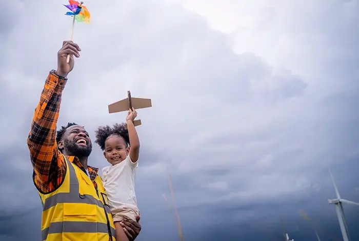 IMG: A father holding his daughter with wind turbines in the background, symbolizing a just renewable energy future for upcoming generations in A FutuRE in Progress.